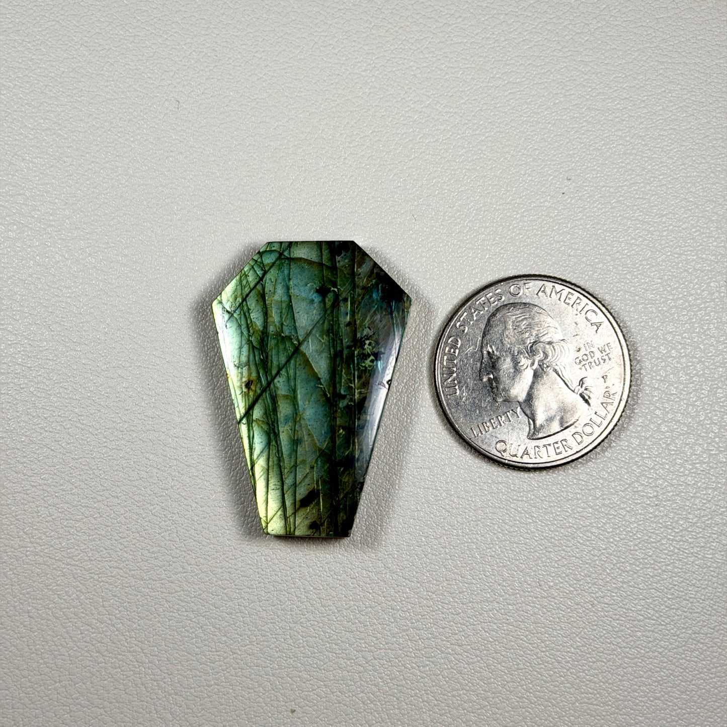 coffin shaped Labradorite stone next to a quarter on a gray background