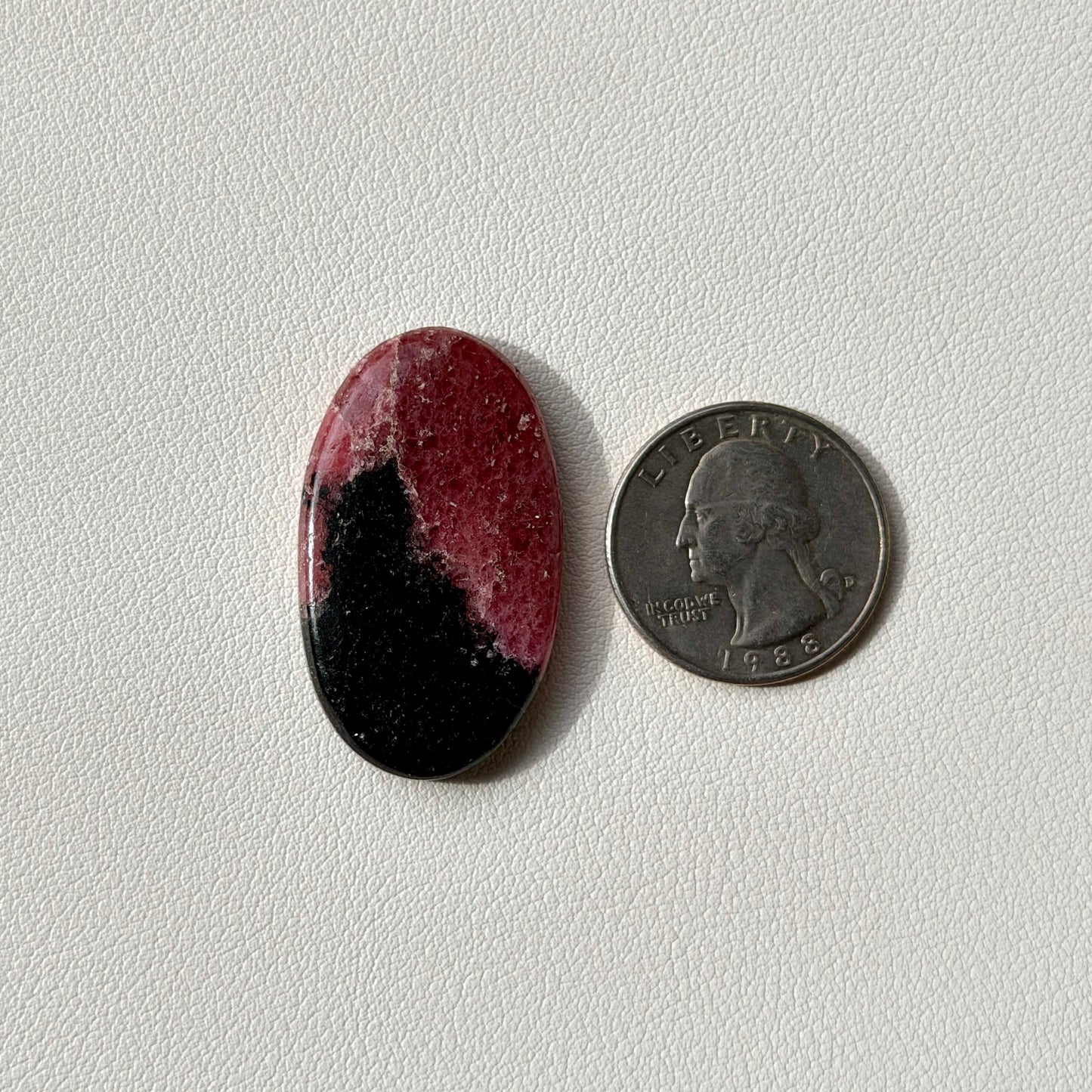 Oval-shaped gemstone with black and pink colors next to a U.S. quarter on a light gray background