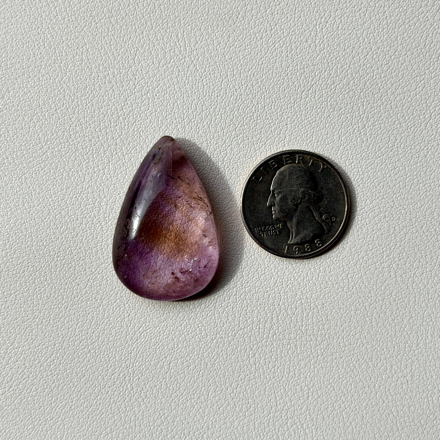 Purple teardrop-shaped stone next to a U.S. quarter on a light gray background