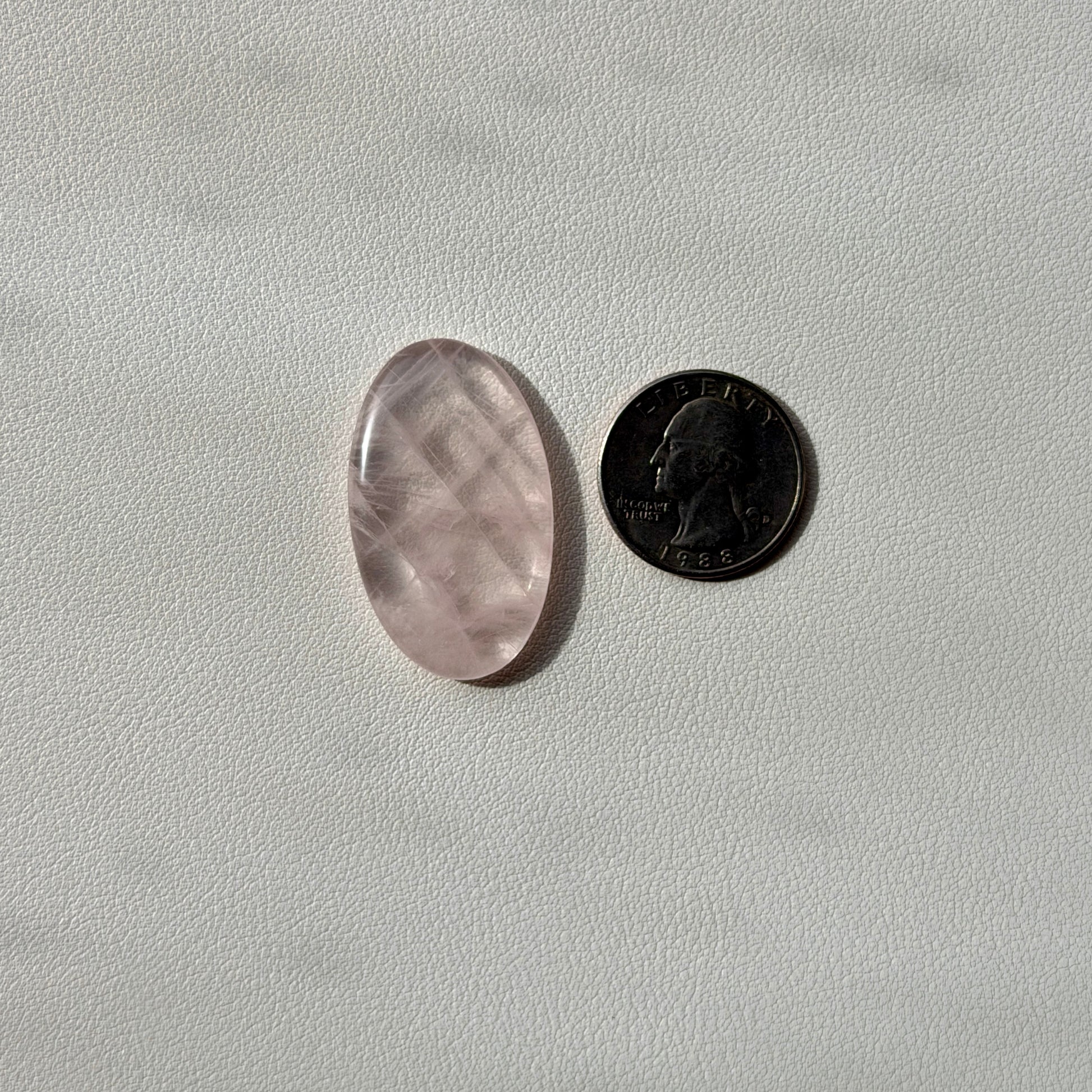 Oval-shaped rose quartz crystal next to a U.S. quarter on a light gray background