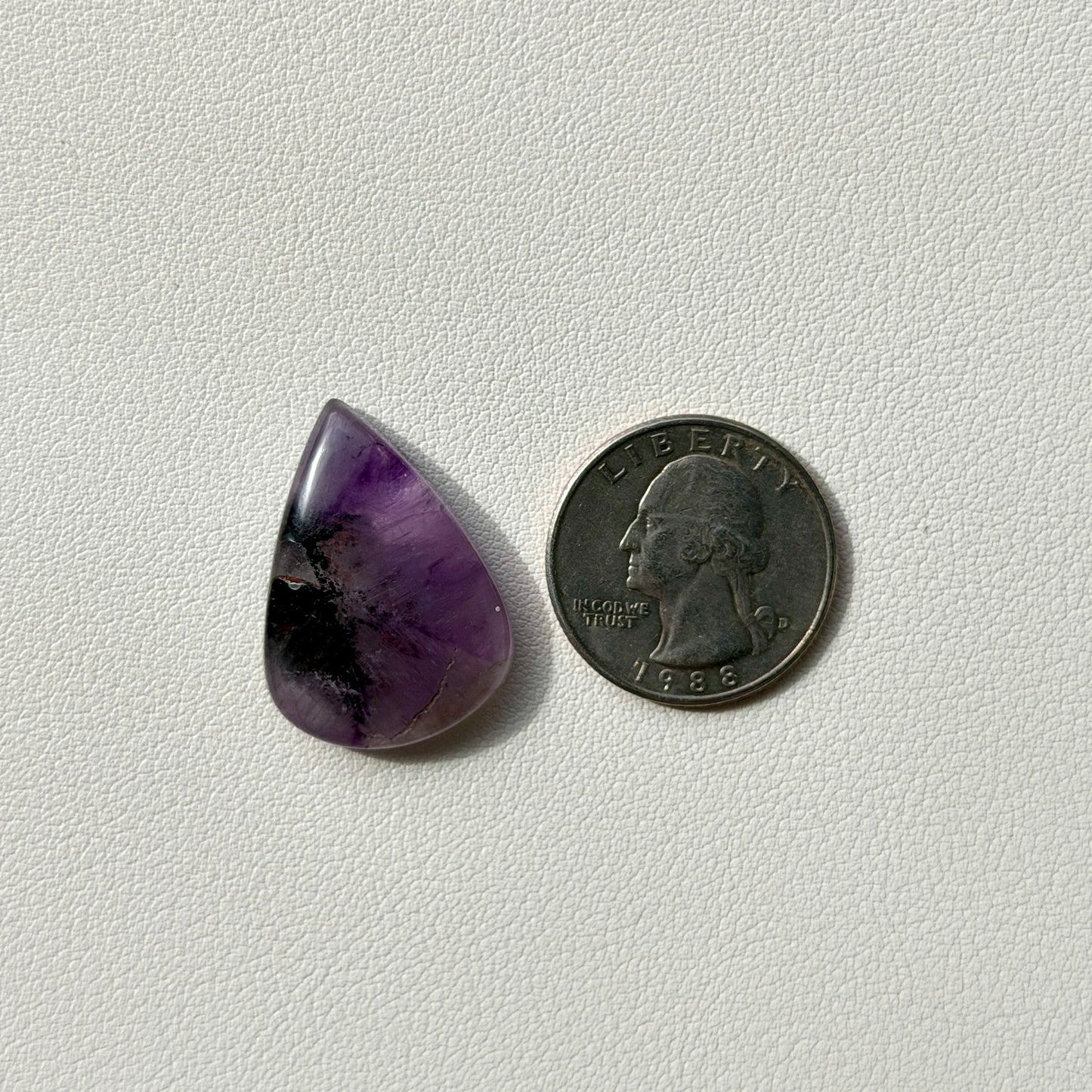 Teardrop-shaped purple stone next to a U.S. quarter on a light gray background