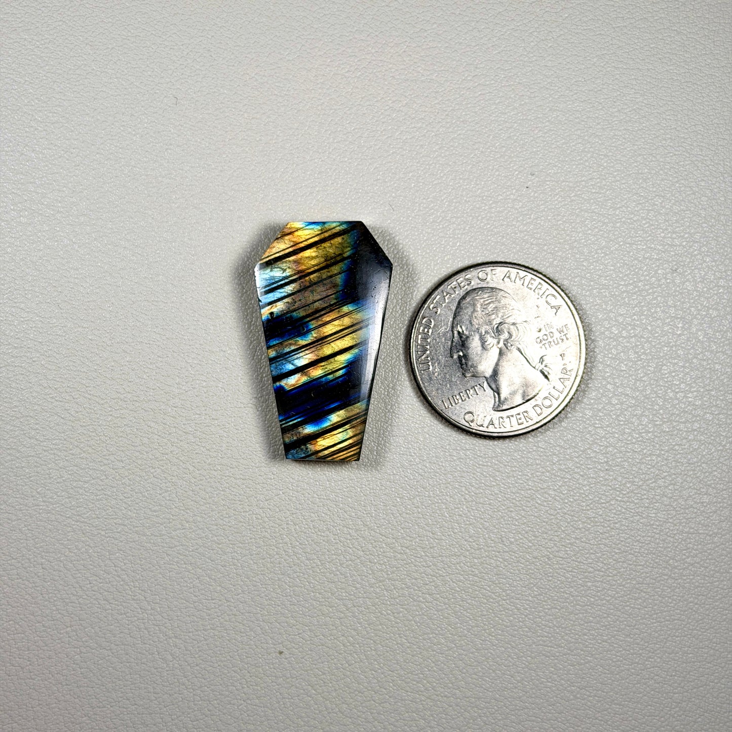 coffin shaped labradorite  stone next to a quarter on a gray background